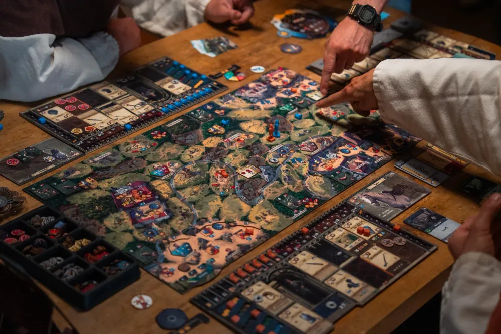 A retail product shot of the Kingdom Come: Deliverance board game, showing the boxed set with the fold-out map board, player boards, cards, and stacks of wooden and cardboard tokens.