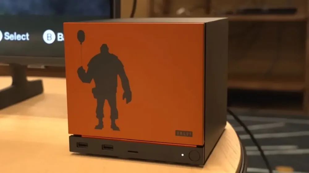 A Steam Machine with an orange Team Fortress 2 Heavy face plate sitting beside a monitor showing the SteamOS interface.
